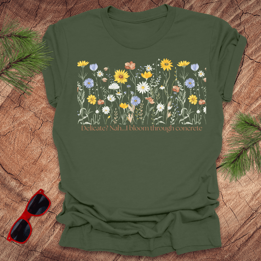 Green t-shirt with floral design and text "delicate? nah. i bloom through concrete.' on a wooden surface.