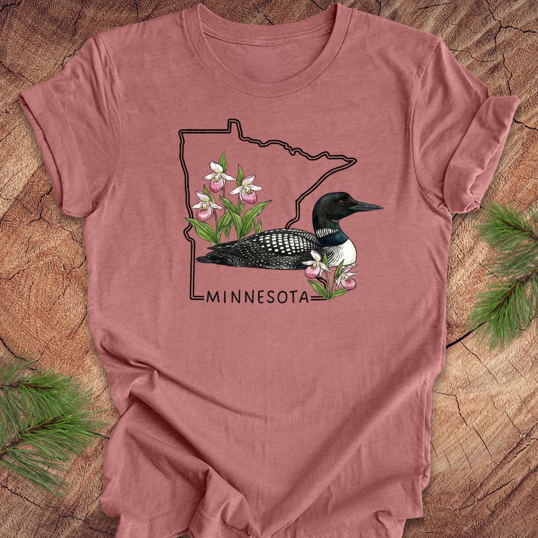 A dusty rose short-sleeve t-shirt laid flat on a rustic wood background with pine branches in the corners. The shirt features a graphic of a loon inside the outline of Minnesota, decorated with pink and white wildflowers, and the word 'MINNESOTA' printed below.