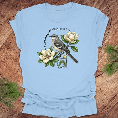 Baby blue t-shirt with the outline of Mississippi with state bird, mockingbird and flower, magnolia, on a wooden surface