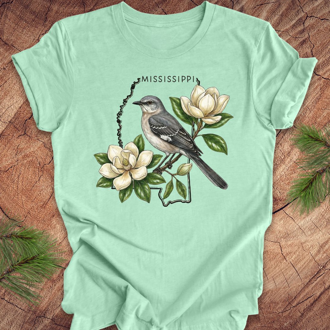 Mint green t-shirt with the outline of Mississippi with state bird, mockingbird and flower, magnolia, on a wooden surface