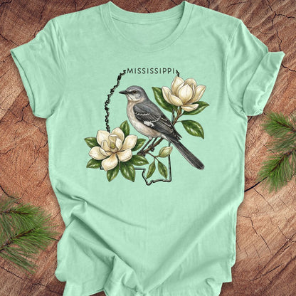 Mint green t-shirt with the outline of Mississippi with state bird, mockingbird and flower, magnolia, on a wooden surface