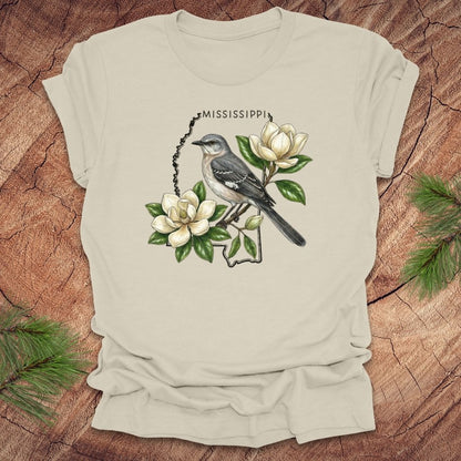 Beige t-shirt with the outline of Mississippi with state bird, mockingbird and flower, magnolia, on a wooden surface