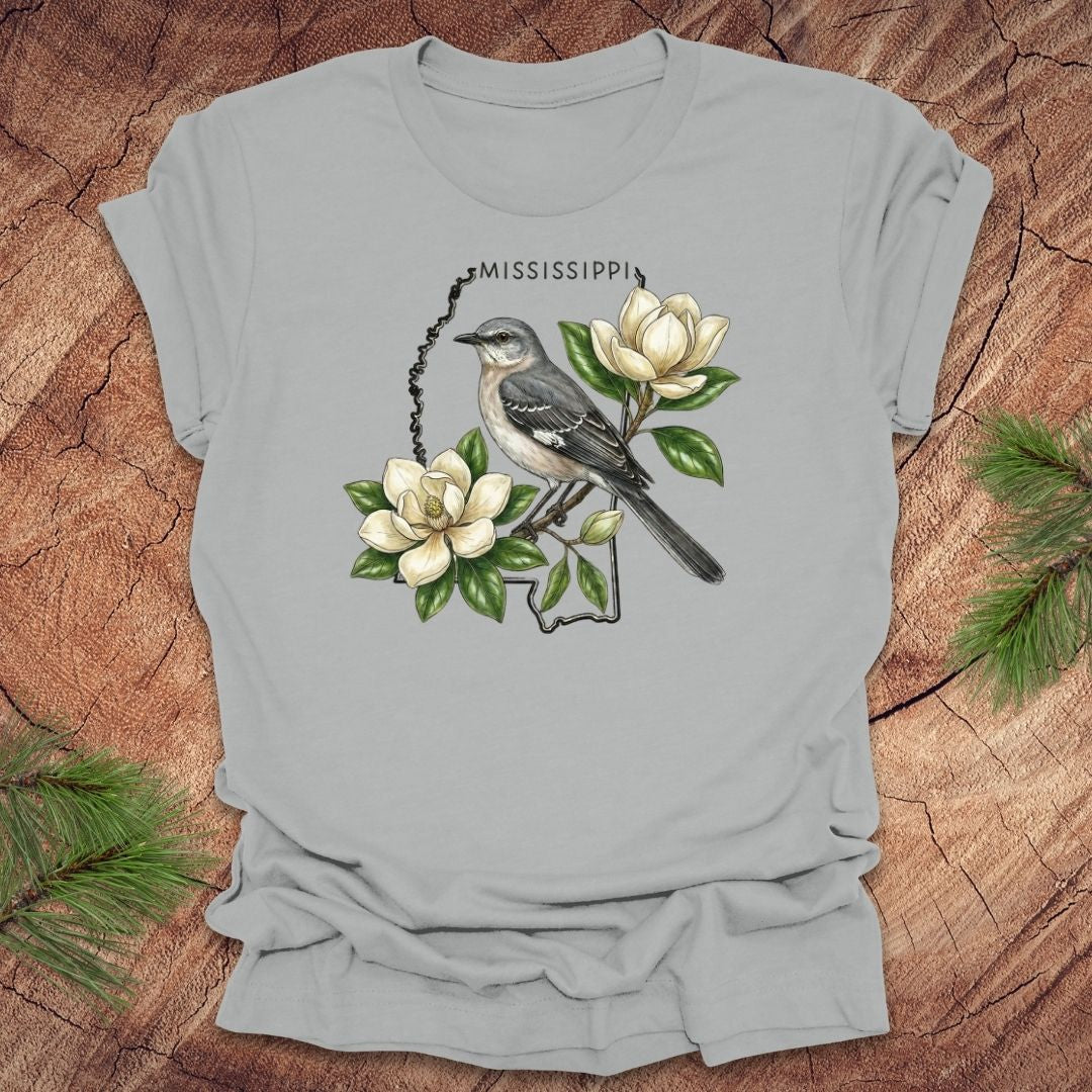 Silver t-shirt with the outline of Mississippi with state bird, mockingbird and flower, magnolia, on a wooden surface