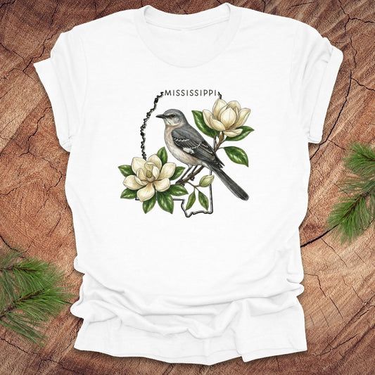 White t-shirt with the outline of Mississippi with state bird, mockingbird and flower, magnolia, on a wooden surface