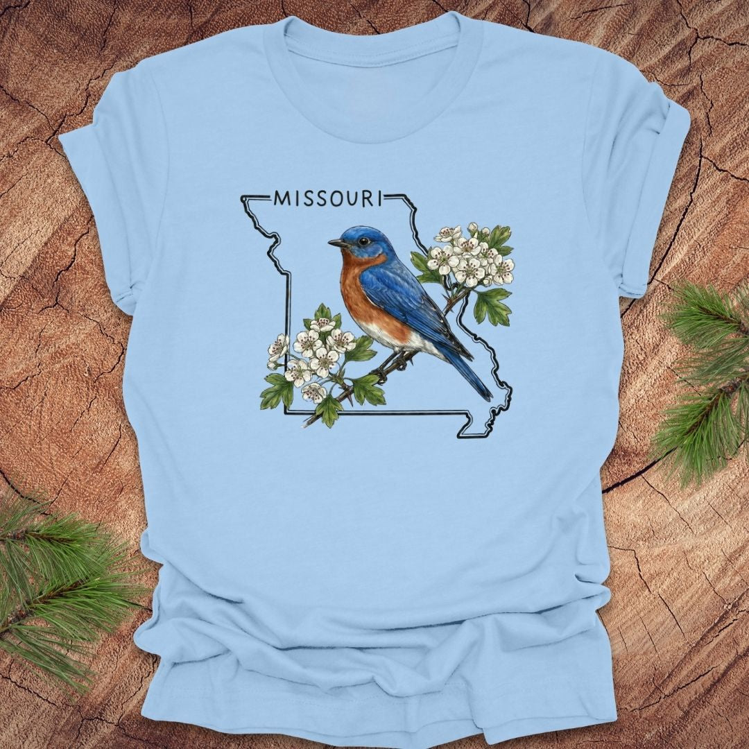 Light blue t-shirt with a Missouri state outline, bluebird, and white flowers on a wooden surface.