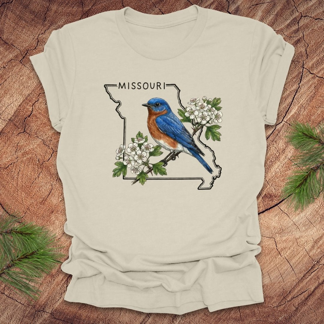 Beige t-shirt with a Missouri state outline and bluebird design on a wooden surface.