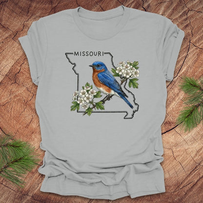 Gray t-shirt with a bluebird and white flowers design on a Missouri map, placed on a wooden surface.