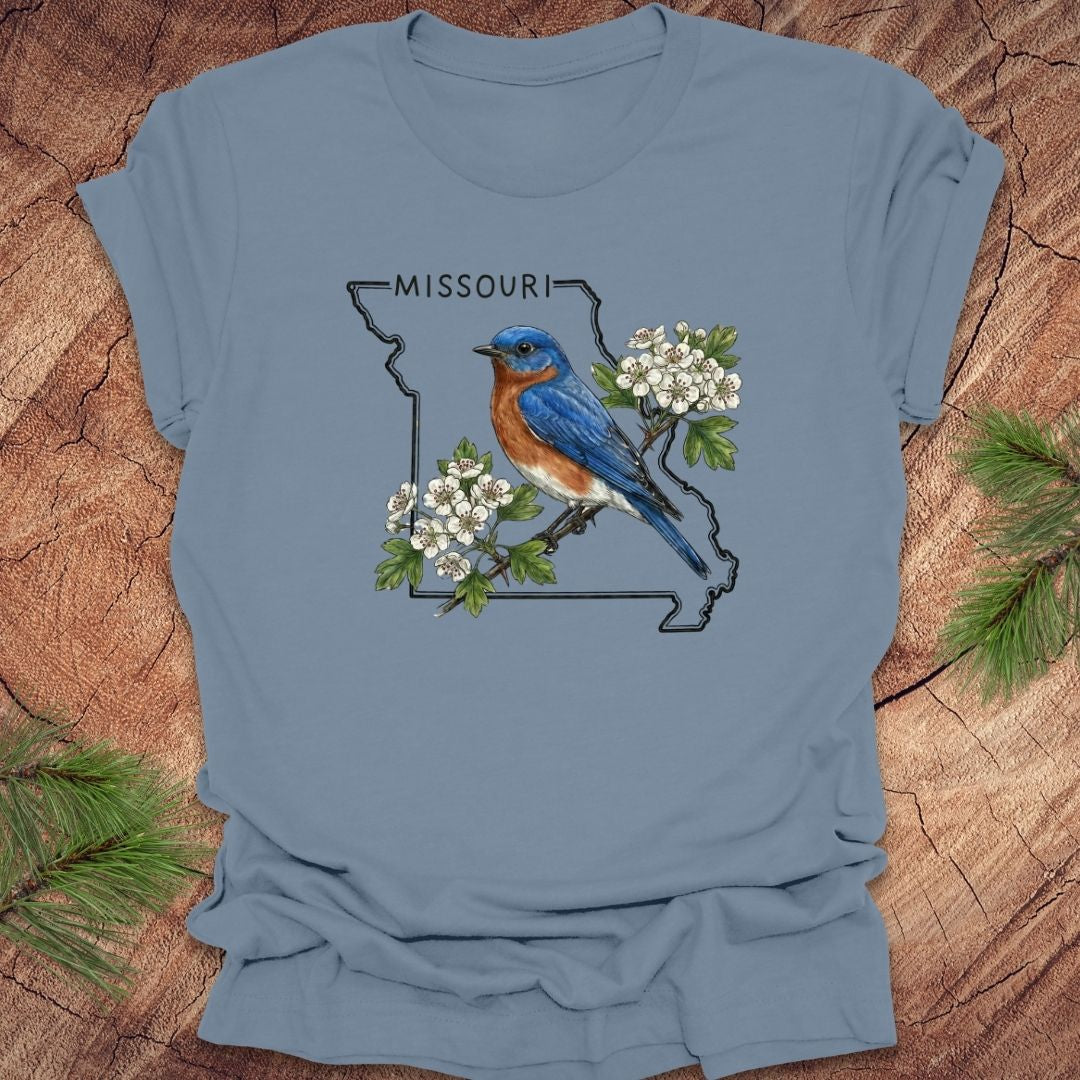 Blue t-shirt with a Missouri state outline, bluebird, and flowers on a wooden surface.