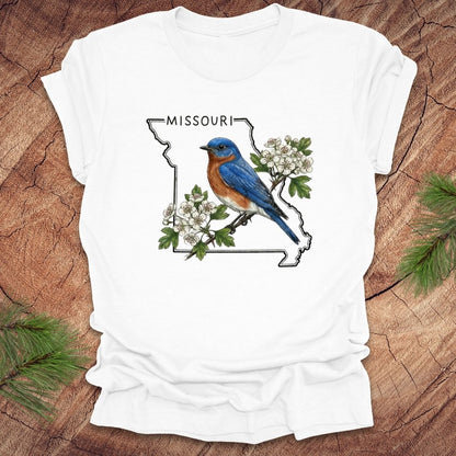 White t-shirt with a bluebird and Missouri state outline design on a wooden surface.