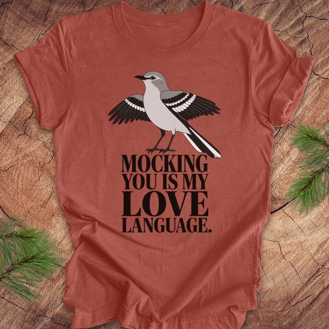 A rust-colored short-sleeve t-shirt with a printed graphic of a mocking bird and the text 'MOCKING YOU IS MY LOVE LANGUAGE.' laid flat on a wood surface with pine branches in the corners.