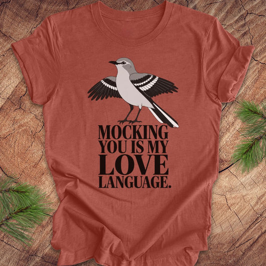 A rust-colored short-sleeve t-shirt with a printed graphic of a mocking bird and the text 'MOCKING YOU IS MY LOVE LANGUAGE.' laid flat on a wood surface with pine branches in the corners.