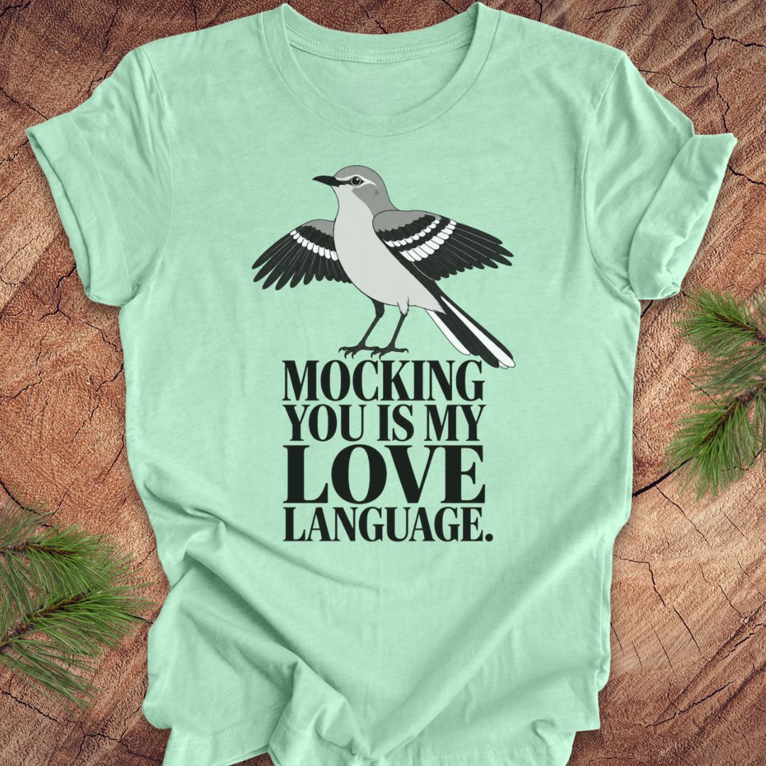 A mint green short-sleeve t-shirt laid flat on a rustic wood background with pine branches in the corners. The shirt features a graphic of a gray and black mocking bird above the text 'MOCKING YOU IS MY LOVE LANGUAGE.