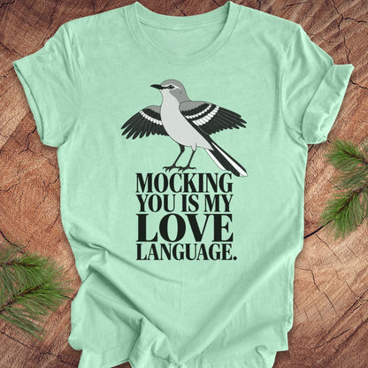 A mint green short-sleeve t-shirt laid flat on a rustic wood background with pine branches in the corners. The shirt features a graphic of a gray and black mocking bird above the text 'MOCKING YOU IS MY LOVE LANGUAGE.
