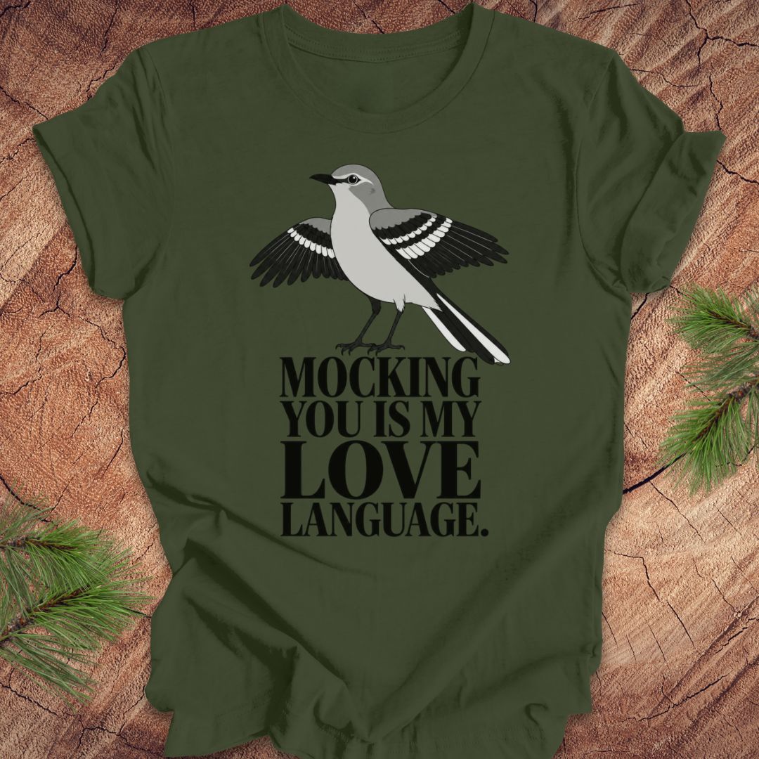 An olive green short-sleeve t-shirt with a graphic of a gray and black mockingbird and the text 'MOCKING YOU IS MY LOVE LANGUAGE.' printed on the front, displayed flat on a rustic wood background with pine needles.