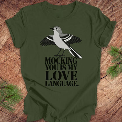 An olive green short-sleeve t-shirt with a graphic of a gray and black mockingbird and the text 'MOCKING YOU IS MY LOVE LANGUAGE.' printed on the front, displayed flat on a rustic wood background with pine needles.