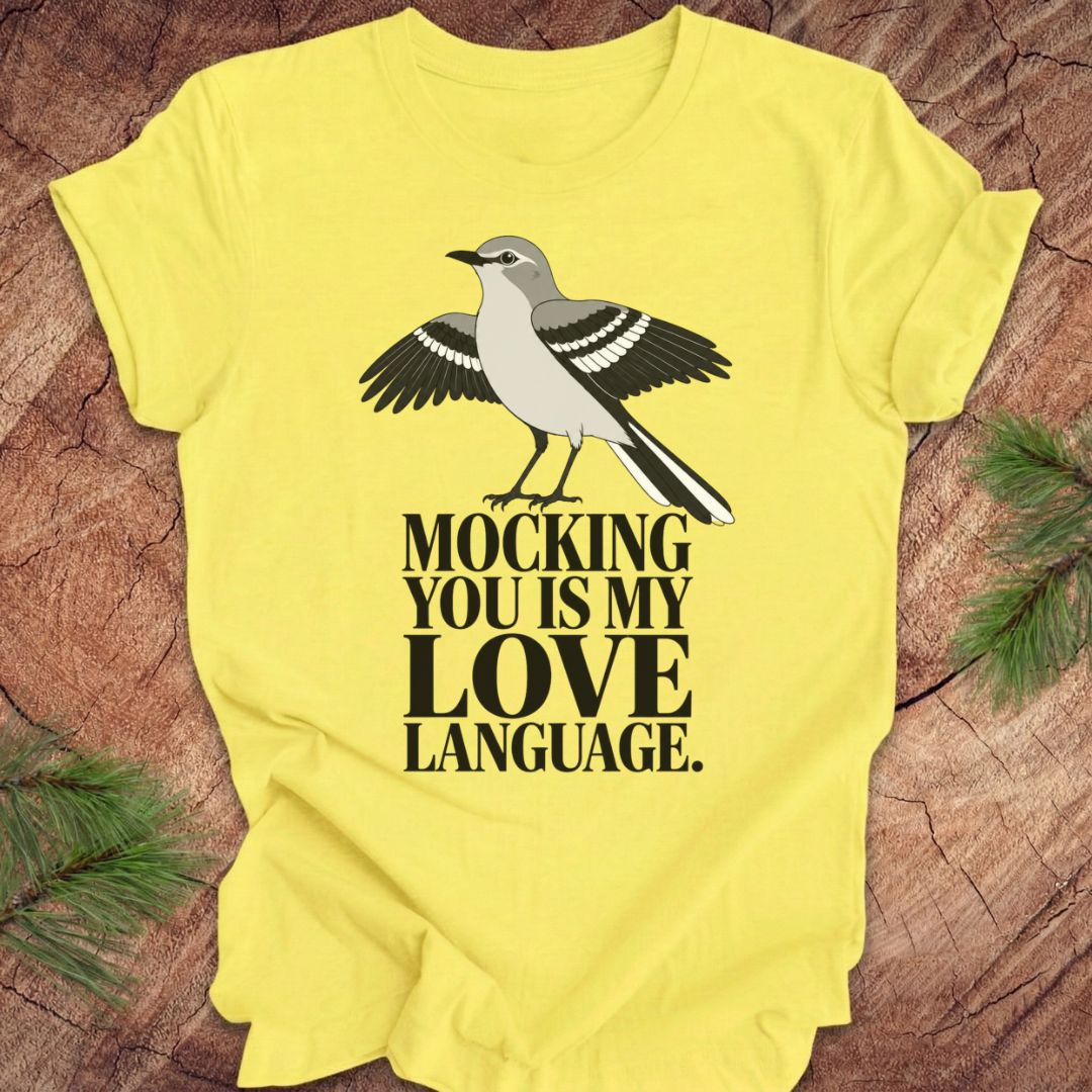 A bright yellow short-sleeve t-shirt laid flat on a rustic wood surface with pine branches at the corners. The shirt features a printed graphic of a gray and black bird above the bold black text, 'MOCKING YOU IS MY LOVE LANGUAGE.