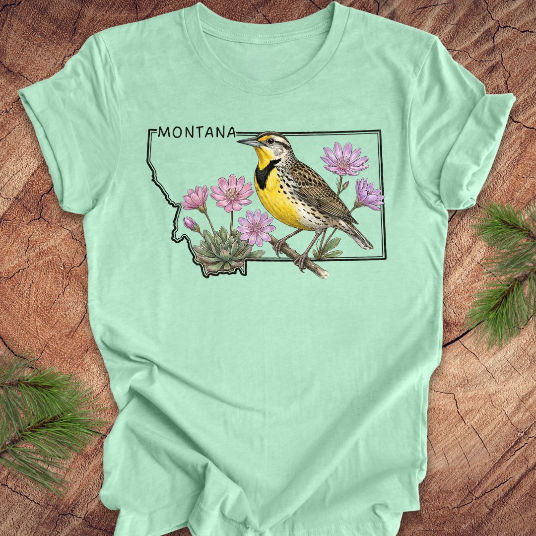 Mint green t-shirt with the state outline of Montana with a graphic of a western meadowlark and bitteroot, on a wooden surface