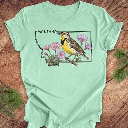 Mint green t-shirt with the state outline of Montana with a graphic of a western meadowlark and bitteroot, on a wooden surface
