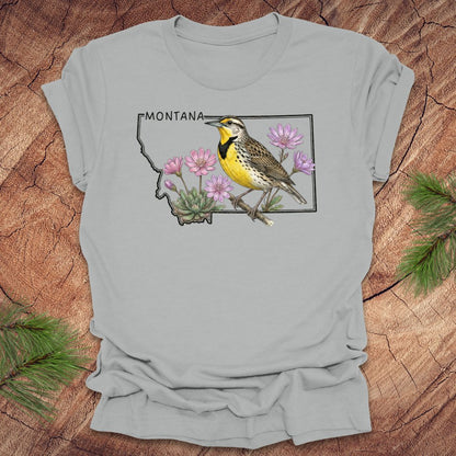 Silver t-shirt with the state outline of Montana with a graphic of a western meadowlark and bitteroot, on a wooden surface
