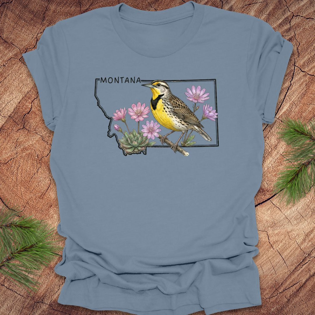 Steel blue t-shirt with the state outline of Montana with a graphic of a western meadowlark and bitteroot, on a wooden surface