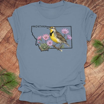 Steel blue t-shirt with the state outline of Montana with a graphic of a western meadowlark and bitteroot, on a wooden surface