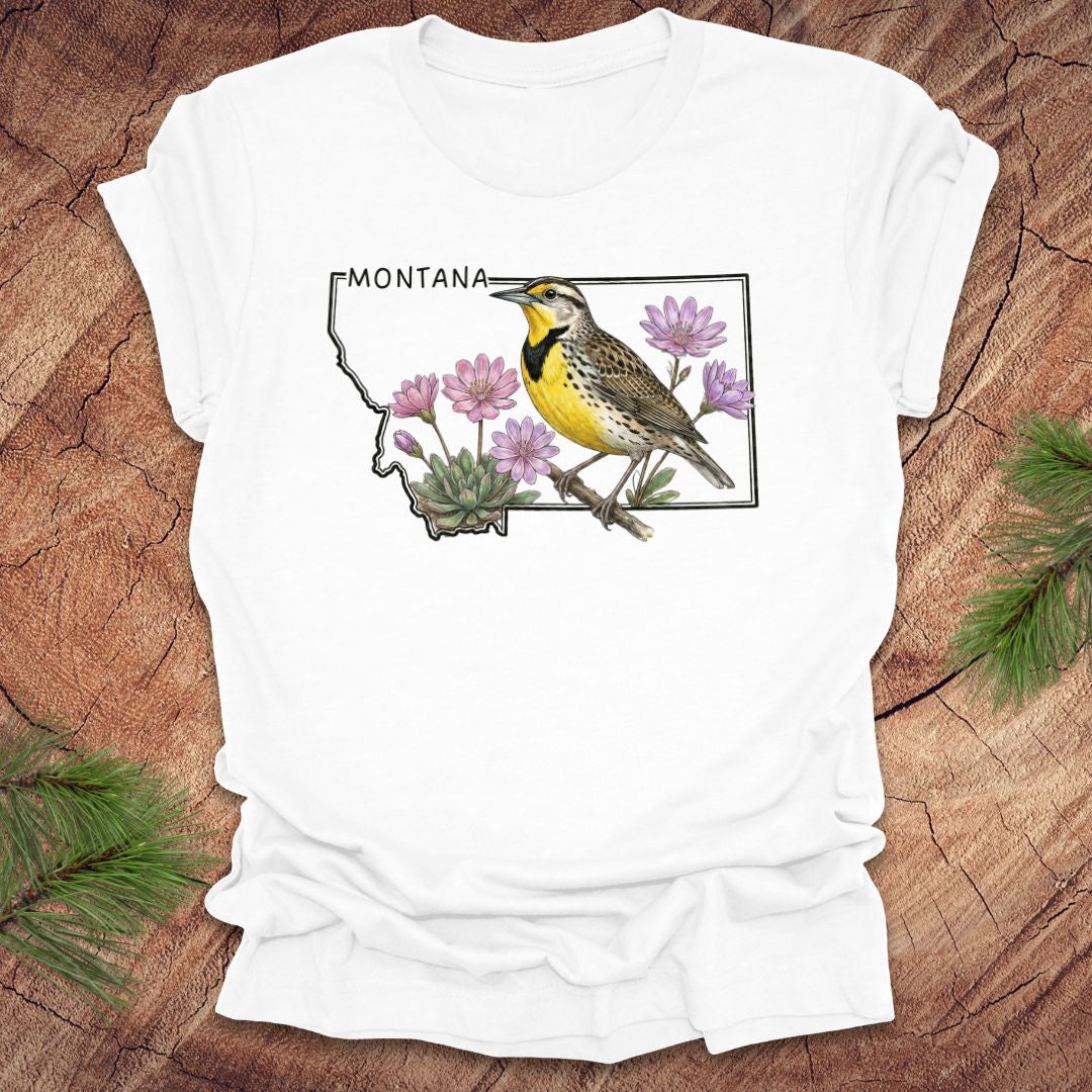 White t-shirt with the state outline of Montana with a graphic of a western meadowlark and bitteroot, on a wooden surface