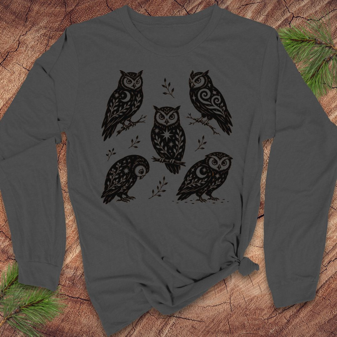 Asphalt gray long-sleeve t-shirt with black folk-art owls decorated with crescent moons, stars, and vines, laid flat on a rustic wood background with pine branches.