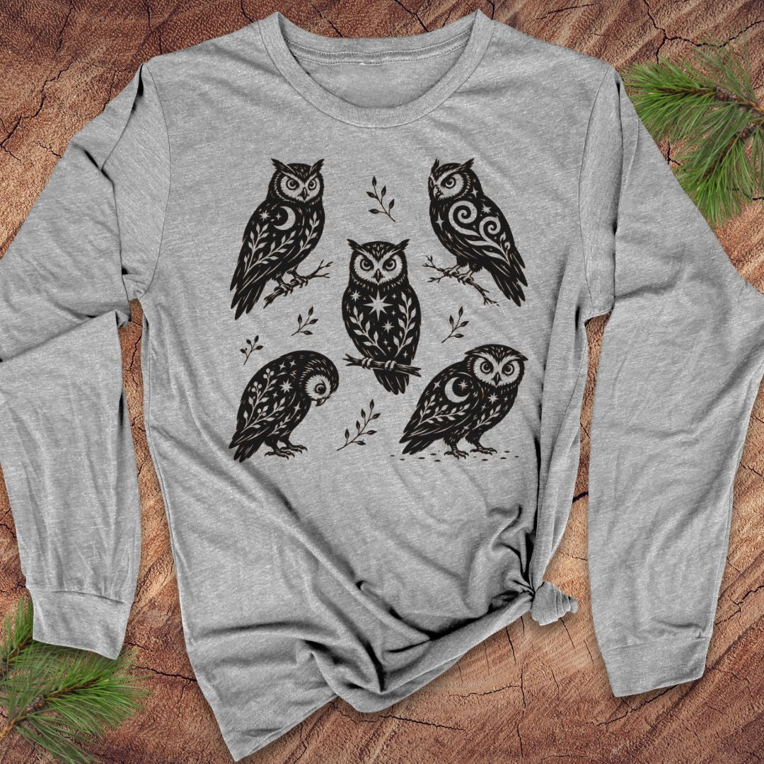Athletic heather gray long-sleeve t-shirt with black folk-art owls decorated with crescent moons, stars, and vines, laid flat on a rustic wood background with pine branches.