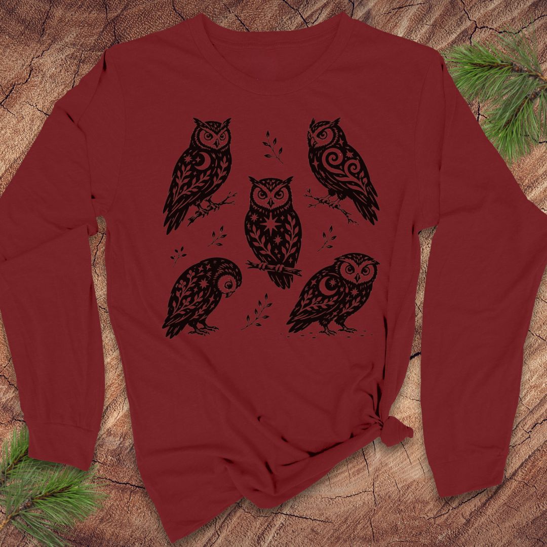 Cardinal red long-sleeve t-shirt with black folk-art owls decorated with crescent moons, stars, and vines, laid flat on a rustic wood background with pine branches.