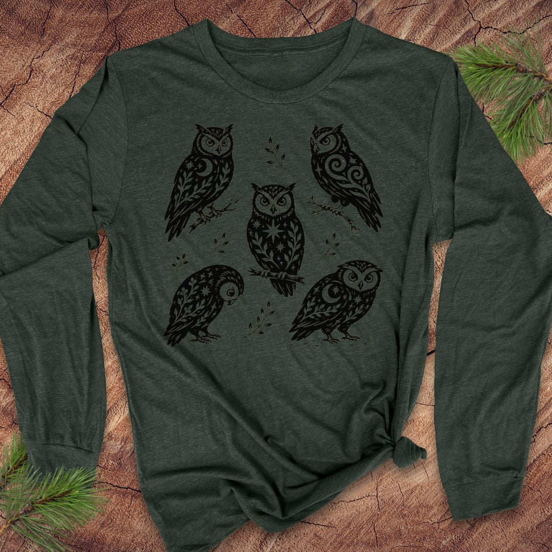 Heather forest green long-sleeve t-shirt with black folk-art owls decorated with crescent moons, stars, and vines, laid flat on a rustic wood background with pine branches.