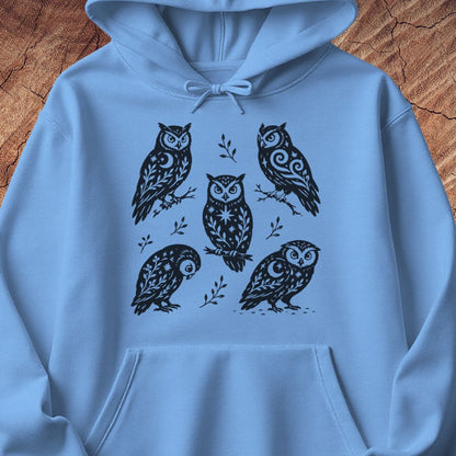 Carolina blue hoodie with black folk-art owls decorated with crescent moons, stars, and vines, laid flat on a rustic wood background.