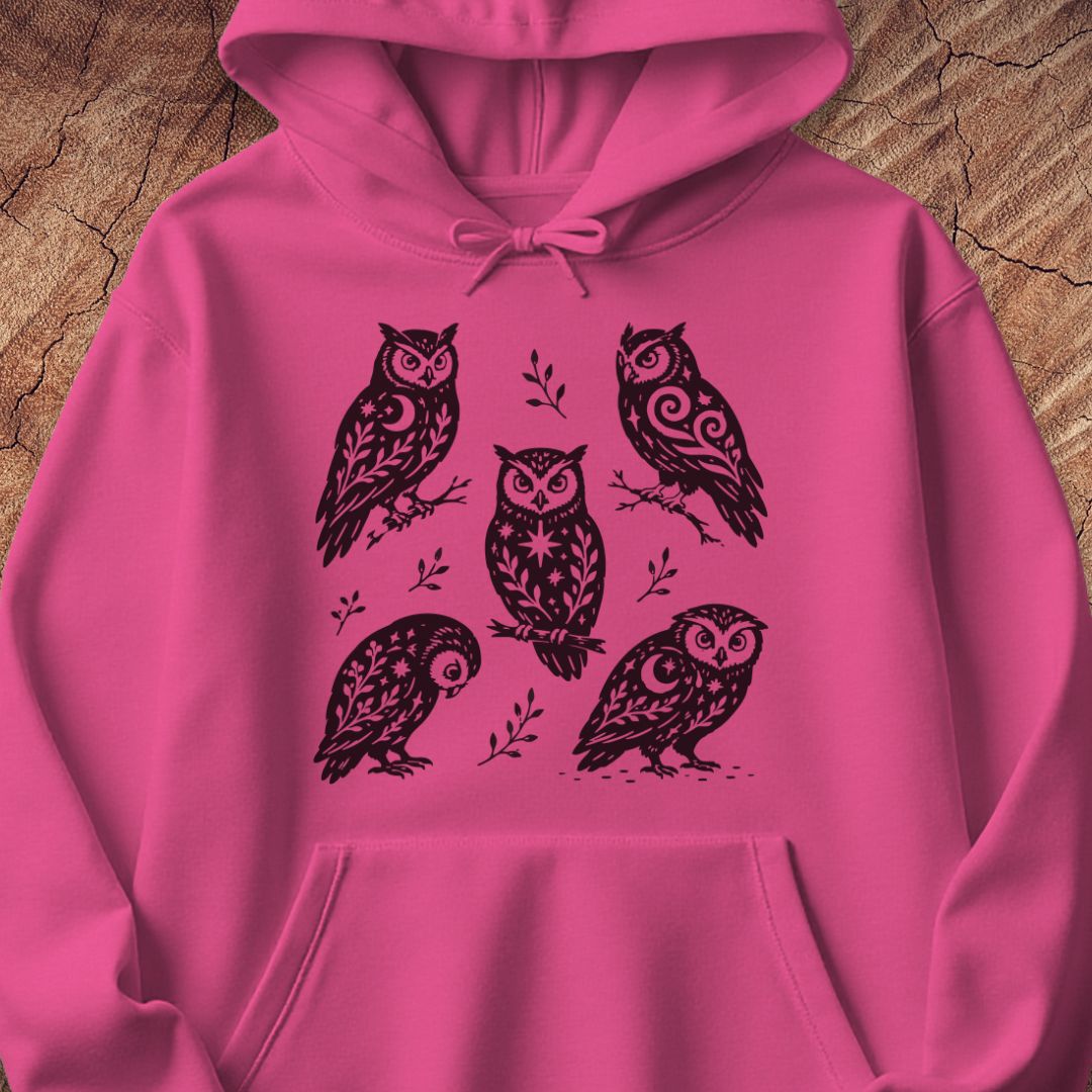 Heliconia pink hoodie with black folk-art owls decorated with crescent moons, stars, and vines, laid flat on a rustic wood background.