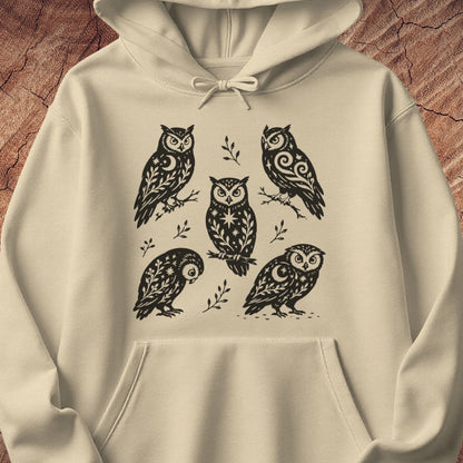 Sand beige hoodie with black folk-art owls decorated with crescent moons, stars, and vines, laid flat on a rustic wood background.