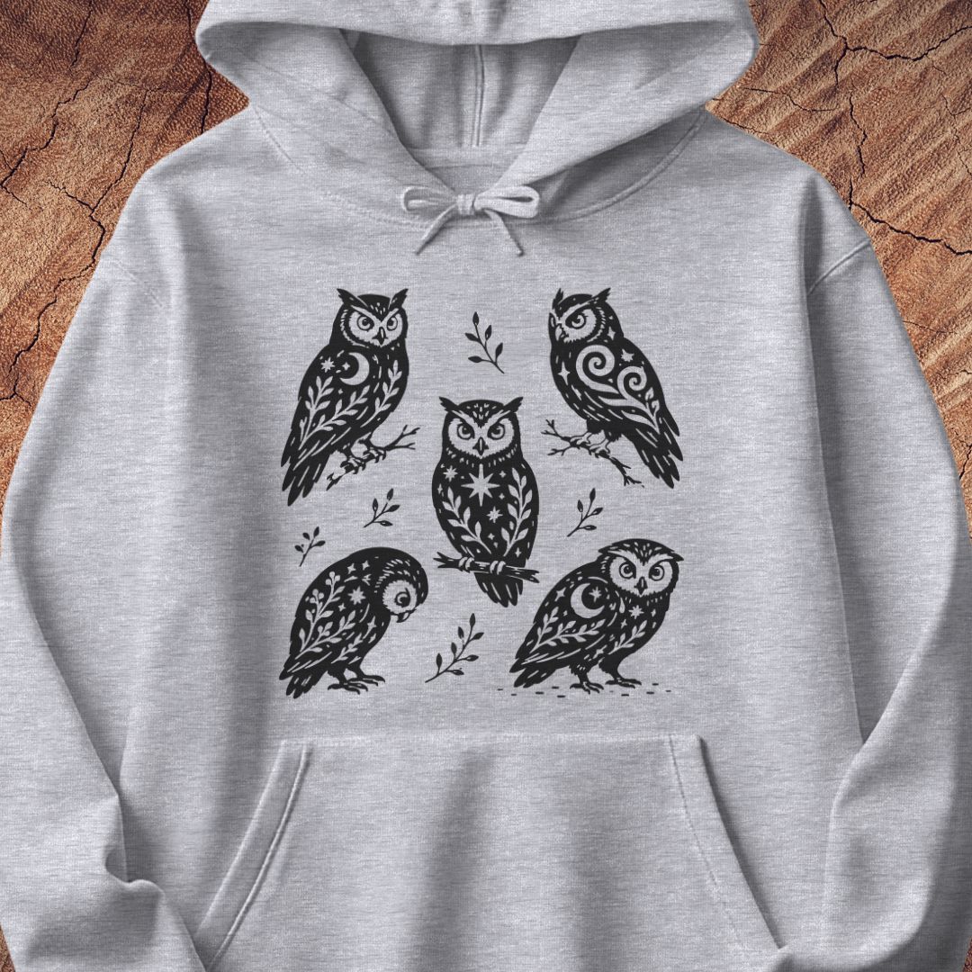 Sport grey hoodie with black folk-art owls decorated with crescent moons, stars, and vines, laid flat on a rustic wood background.