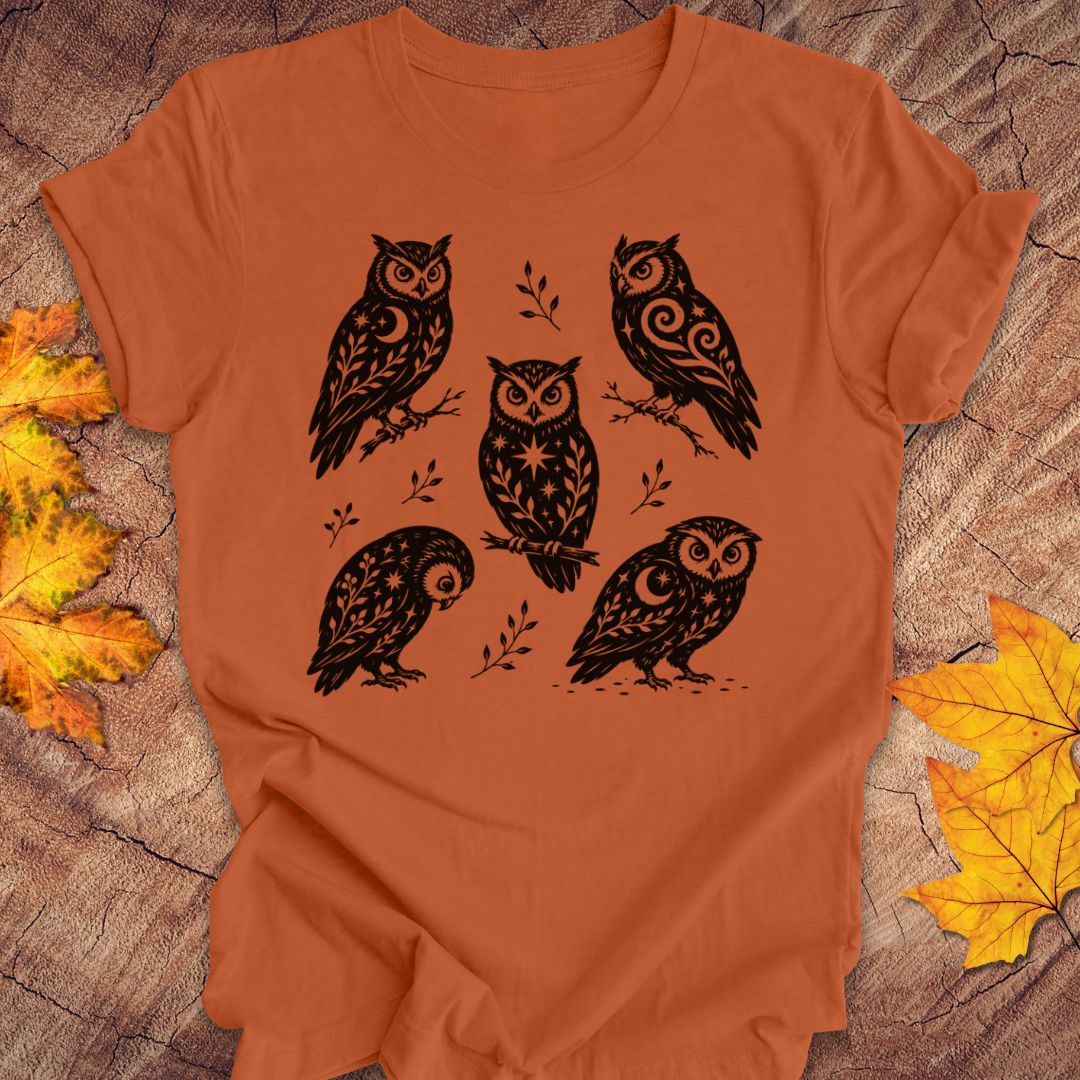 An orange short-sleeve t-shirt with a black graphic print of five owls, laid flat on a rustic wood background with autumn leaves.