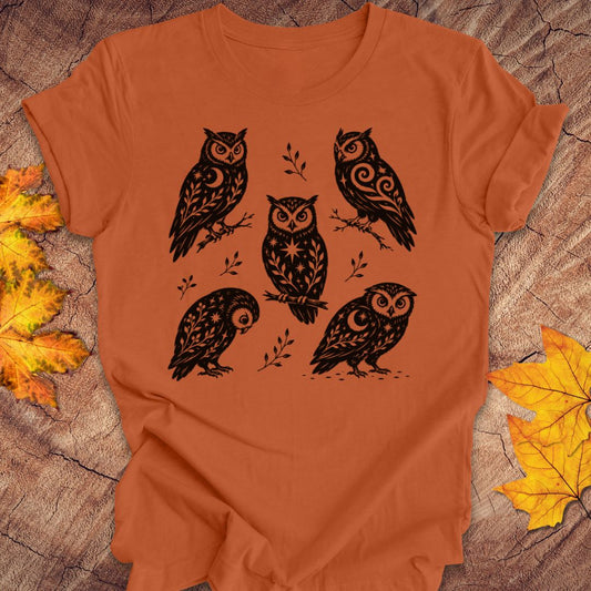An orange short-sleeve t-shirt with a black graphic print of five owls, laid flat on a rustic wood background with autumn leaves.