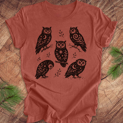 A terracotta-colored short-sleeve t-shirt with a black graphic print of five stylized owls, laid flat on a rustic wood background with pine branches in the corners.