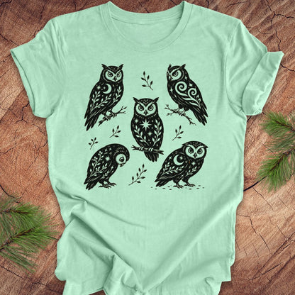 A mint green short-sleeve t-shirt with a black graphic print of five stylized owls, laid flat on a textured brown wood background with pine branches in the corners.