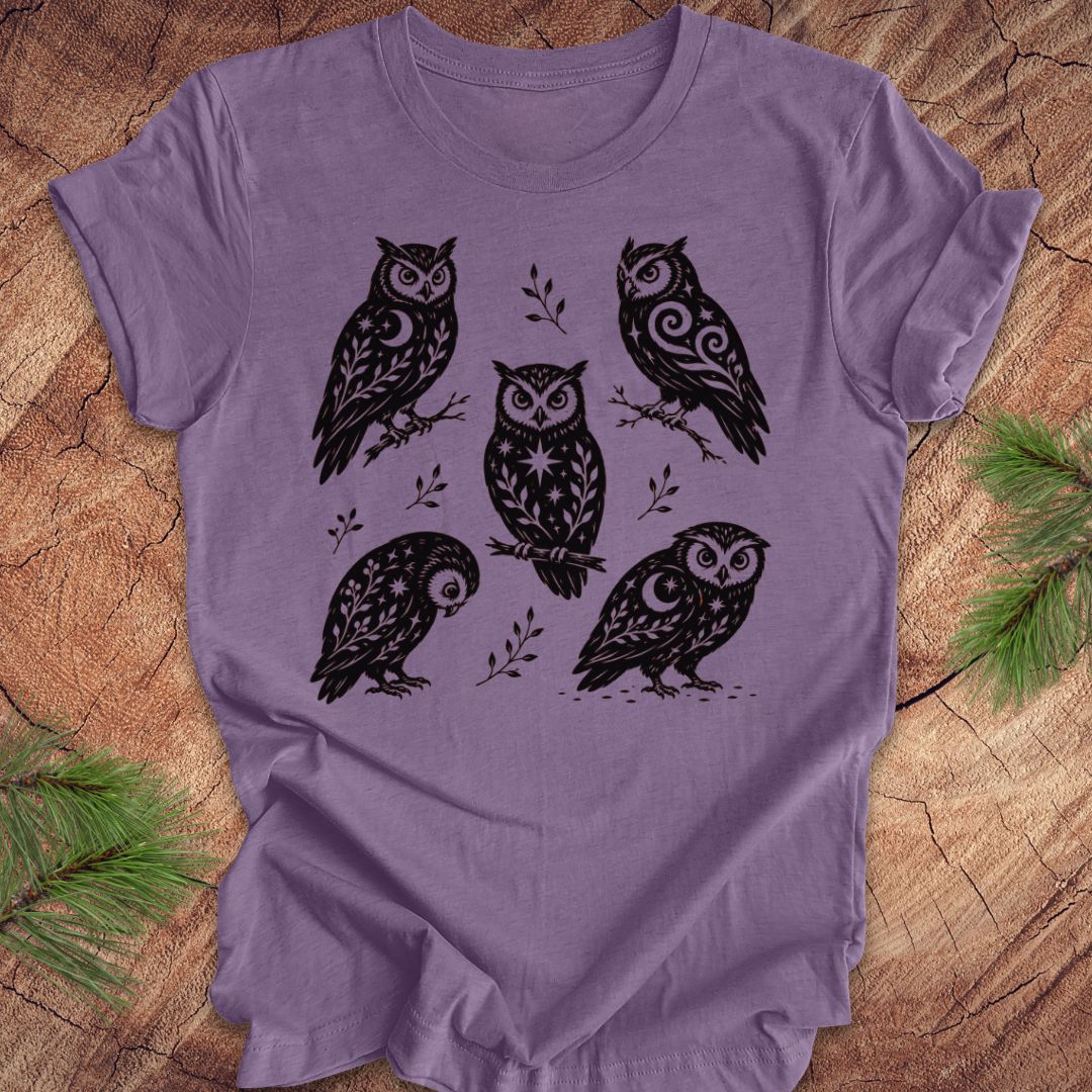 A lavender purple short-sleeve t-shirt with a black graphic print of five stylized owls on branches, laid flat on a rustic wood background with pine needles in the corners.