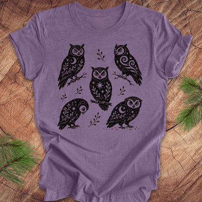 A lavender purple short-sleeve t-shirt with a black graphic print of five stylized owls on branches, laid flat on a rustic wood background with pine needles in the corners.