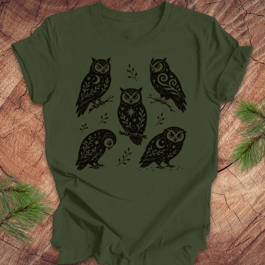 An olive green short-sleeve t-shirt with a black graphic print of five stylized owls, laid flat on a rustic wood background with pine branches in the corners.
