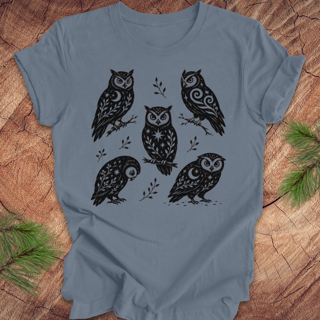 A slate blue short-sleeve t-shirt with a black graphic print of six owls in a folk-art style, laid flat on a rustic wood background with pine branches in the corners.