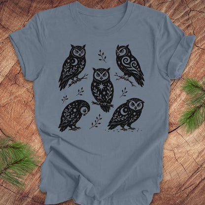A slate blue short-sleeve t-shirt with a black graphic print of six owls in a folk-art style, laid flat on a rustic wood background with pine branches in the corners.