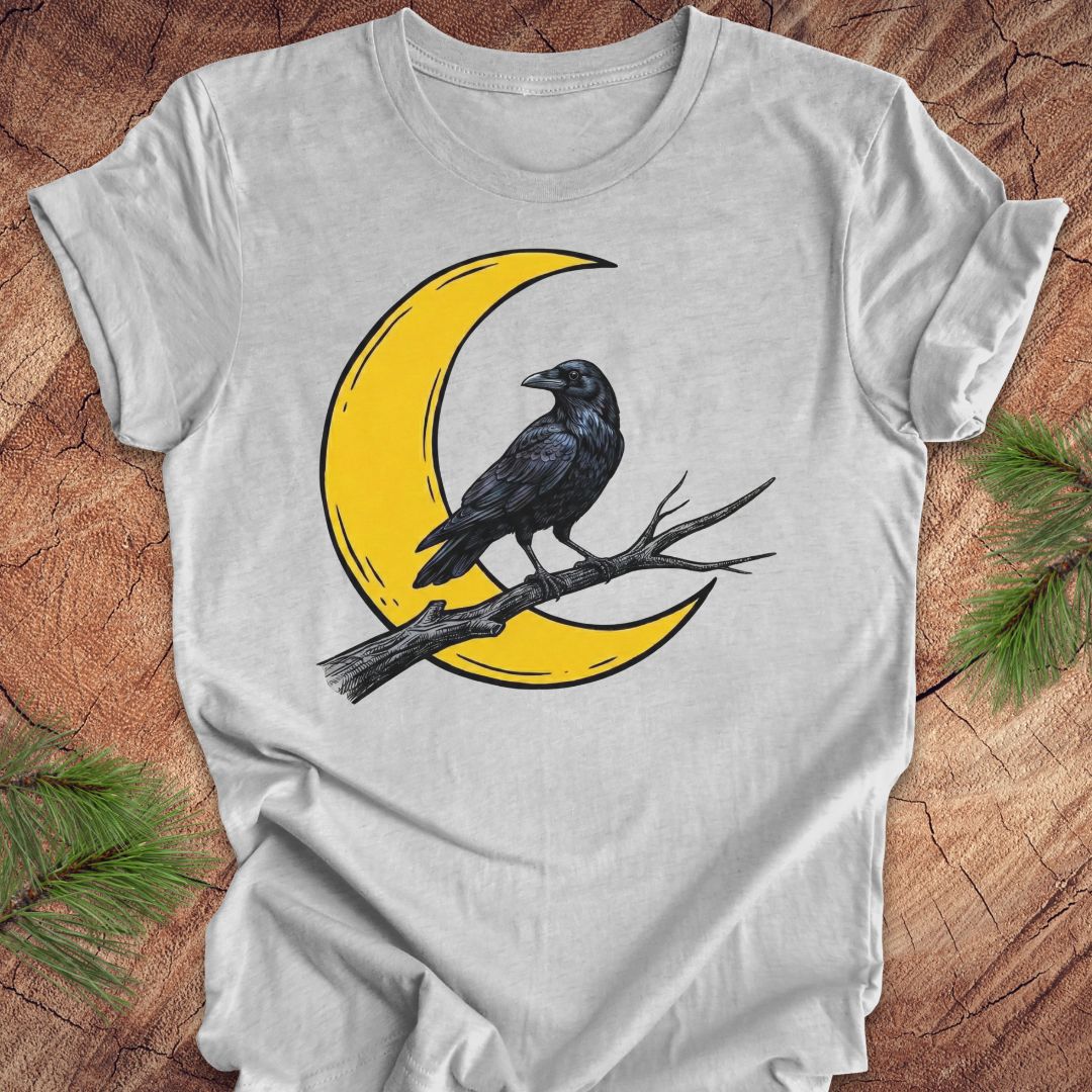 A light gray short-sleeve t-shirt with a graphic of a black raven perched on a yellow crescent moon branch, laid flat on a rustic wood background with pine needles in the corners.