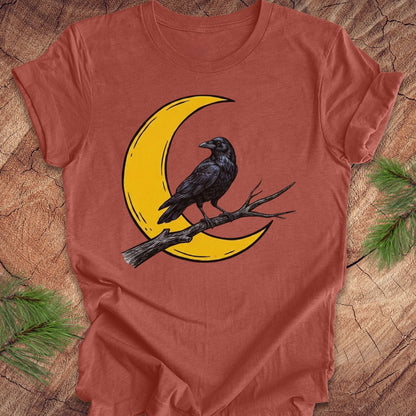A rust-orange short-sleeve t-shirt with a graphic of a black raven perched on a branch in front of a yellow crescent moon, laid flat on a wood surface with pine branches in the corners.