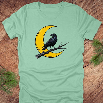 Light green t-shirt with a yellow crescent moon and black crow design on a wooden surface.