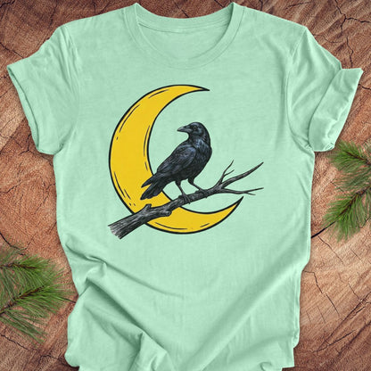 A mint green short-sleeve t-shirt laid flat on a rustic wood background, featuring a graphic of a black raven perched on a yellow crescent moon branch.