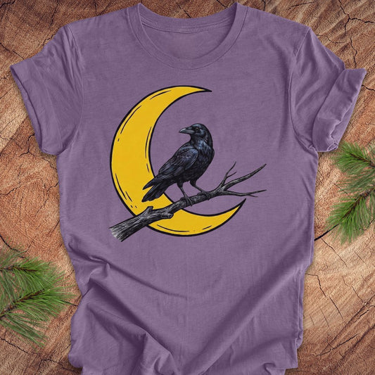 A purple short-sleeve t-shirt laid flat on a rustic wood background with pine branches in the corners. The shirt features a graphic of a black raven perched on a branch in front of a yellow crescent moon.
