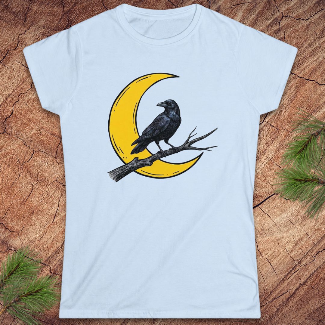 A light blue short-sleeve crewneck t-shirt featuring a graphic of a black raven perched on a branch in front of a yellow crescent moon, laid flat on a rustic wood background with pine needles in the corners.