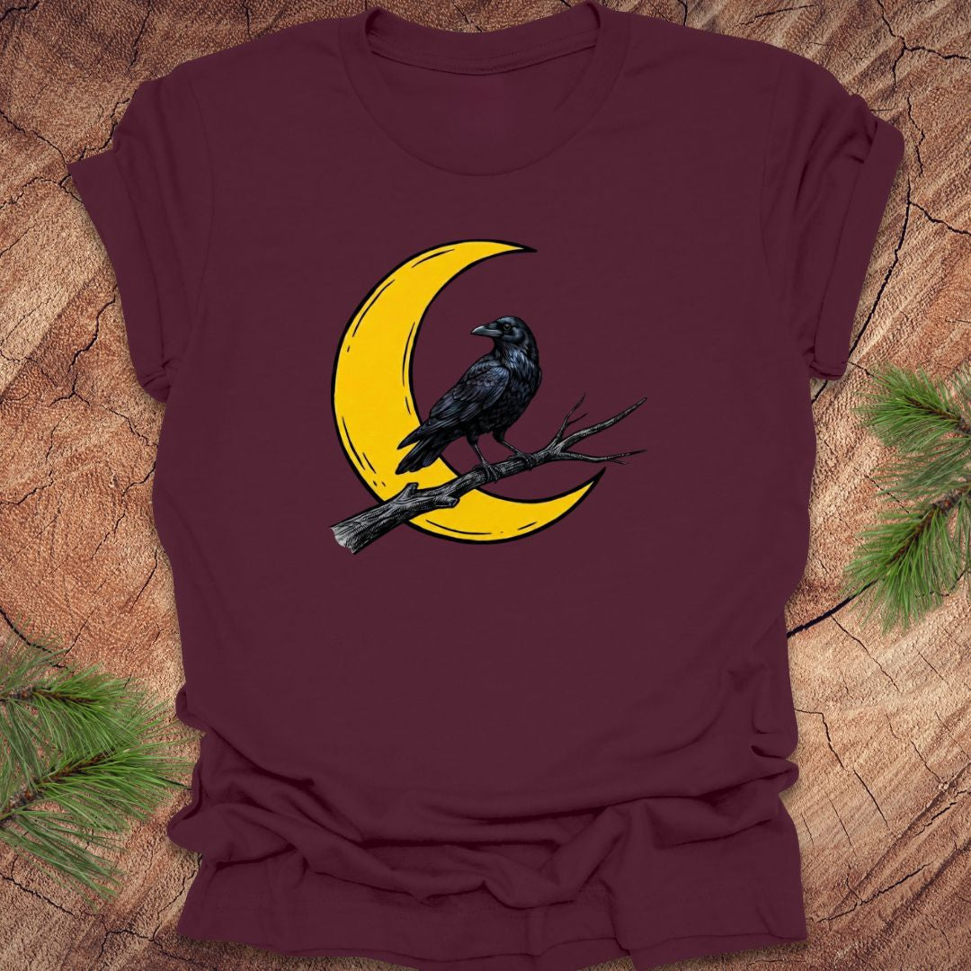 Maroon t-shirt with a yellow crescent moon and black crow design on a wooden surface.